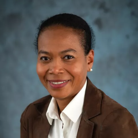 Lepida sits smiling at the camera. She wears a brown blazer over a white shirt.