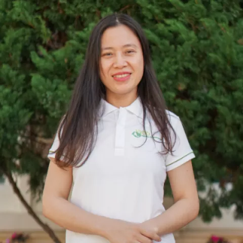 Lieu smiles warmly, wearing a white polo shirt. She stands against a backdrop of lush green foliage.