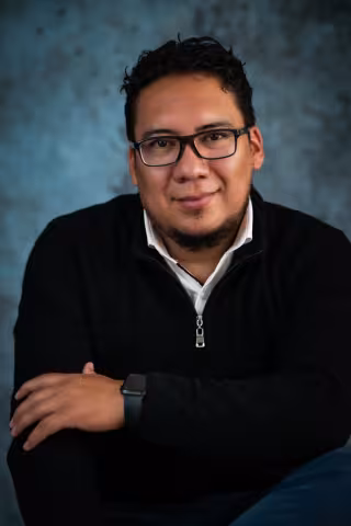 Luis smiles softly at the camera while wearing a black crew neck sweater. 