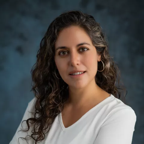 Soledad smiles softly looking into the camera. She wears a white shirt. The background is a mottled blue.