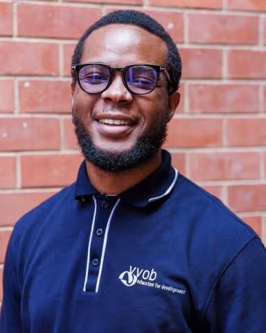 Mawande smiling in a navy polo shirt stands against a brick wall. His expression is warm and friendly.