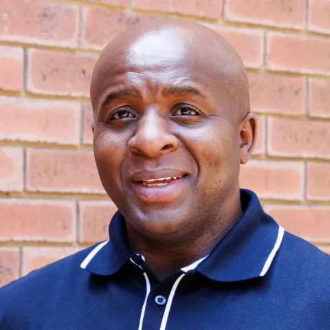 Nkululeko smiling in a navy polo shirt stands against a brick wall. His expression is warm and friendly.