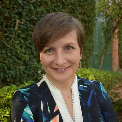 Karolina smiling warmly, standing outdoors against lush green hedges. The setting feels serene and inviting.