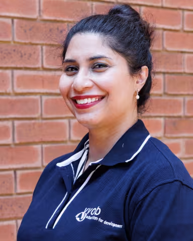 Sahar smiling in a navy polo shirt stands against a brick wall. Her expression is warm and friendly.