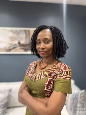 Sarina is wearing a colorful patterned dress and orange necklace, stands confidently with folded arms. The background is a room with blue walls.