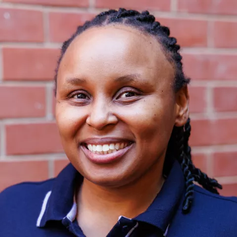 Sisonke smiling in a navy polo shirt stands against a brick wall. Her expression is warm and friendly.