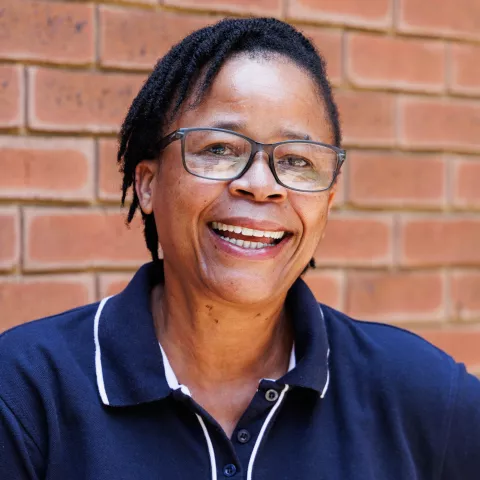 Sylvia smiling in a navy polo shirt stands against a brick wall. Her expression is warm and friendly.