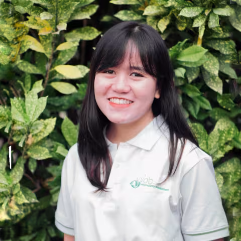 Thao smiles warmly, wearing a white polo shirt. She stands against a backdrop of lush green foliage.