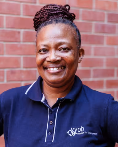 Thobile smiling in a navy polo shirt stands against a brick wall. Her expression is warm and friendly.