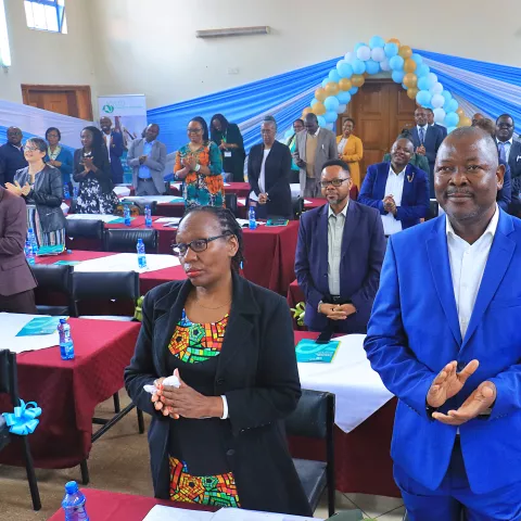A diverse group of people stands applauding in a decorated hall with blue and gold balloons.