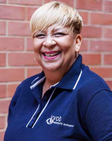 Vanessa smiling in a navy polo shirt stands against a brick wall. Her expression is warm and friendly.