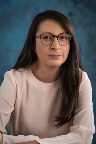 Xiomari smiles softly at the camera. She wears glasses and a beige shirt. The background is a malted blue backdrop.