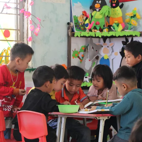 A group of young children sit around a table in a colorful classroom, engaged in an activity.