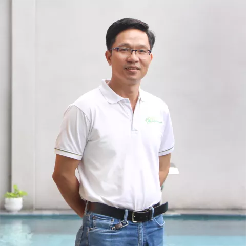 Phuong stands wearing a white VVOB polo shirt and blue jeans and black belt. He is smiling and standing in front of a white background.