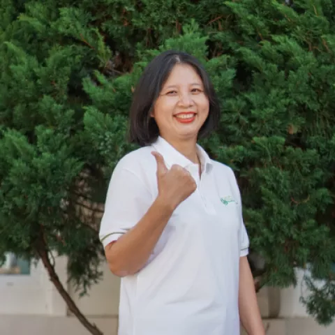 Chau smiles and gives a thumbs up, wearing a white polo shirt, standing in front of lush green foliage. 