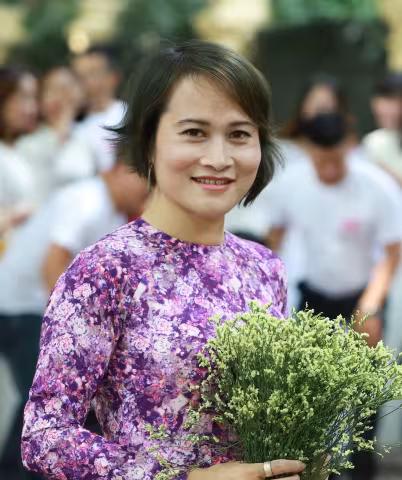 Quyen is wearing purple floral dress smiles warmly, holding a bouquet of delicate white flowers. People in white shirts are blurred in the background.
