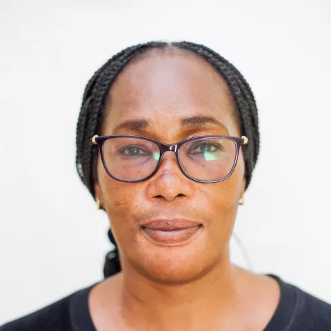 Adonai is wearing glasses and a black top, looking directly at the camera with a calm expression against a light background.