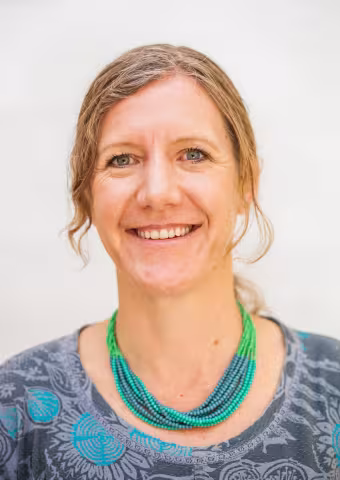 Caroline is wearing a green and blue bead necklace and a gray shirt with a blue floral pattern, against a soft white background.