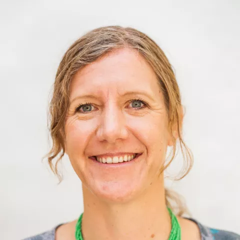 Caroline is wearing a green and blue bead necklace and a gray shirt with a blue floral pattern, against a soft white background.