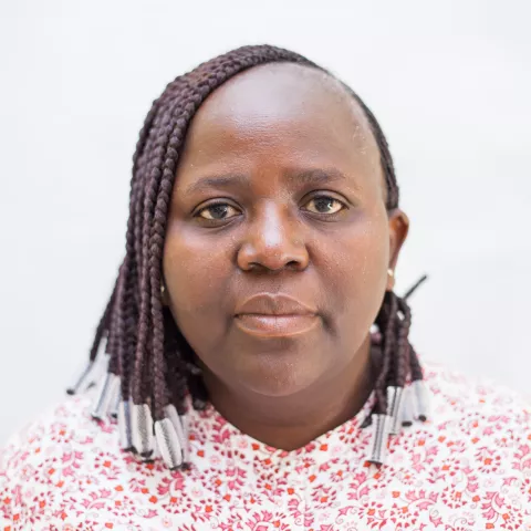 Charity is wearing a red patterned shirt. Her expression is neutral. The background is plain white.  