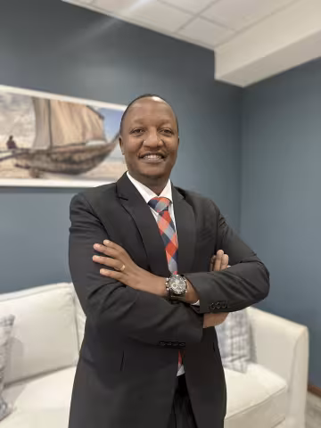 Christopher is wearing a black suit and colorful tie. He stands confidently with arms crossed in an office, a painting of a boat on the wall behind.