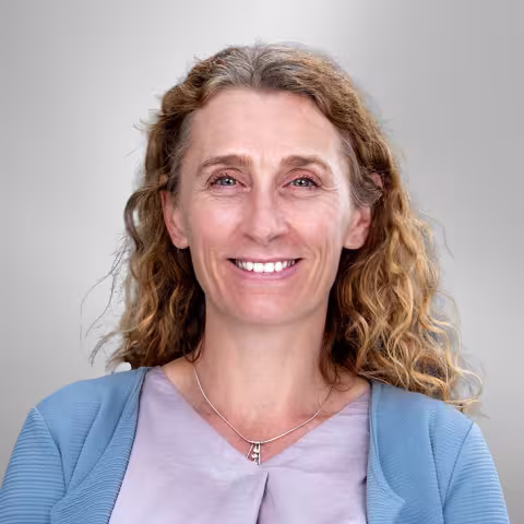 Claire is smiling while wearing a light blue cardigan and silver necklace. She stands against a gray background.