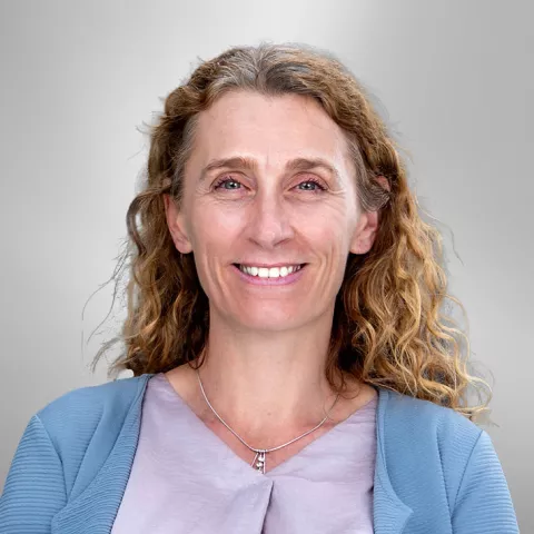 Claire is smiling while wearing a light blue cardigan and silver necklace. She stands against a gray background.