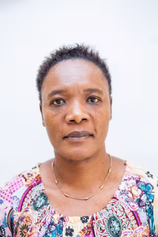 Cynthia looks directly at the camera against a plain white background. She wears a colorful, patterned shirt and a simple gold necklace. Her expression is neutral.