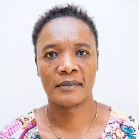 Cynthia looks directly at the camera against a plain white background. She wears a colorful, patterned shirt and a simple gold necklace. Her expression is neutral.