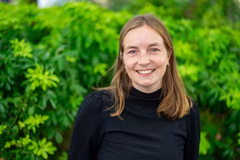 Lisse is wearing a black top and smiles at the camera. The background is green, lush plants.