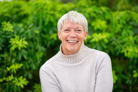 Nancy is wearing a white sweater and smiles. The background is a lush green foliage.