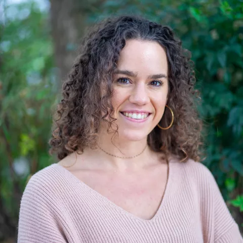Britt is wearing a light sweater and hoop earrings. She stands smiling in front of lush green foliage.