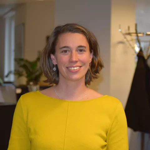 Inge is wearing a yellow top and smiling at the camera. In the background, a modern office is visible.