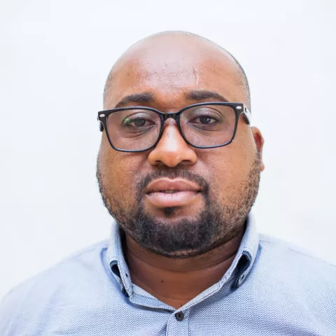 Dean is wearing glasses and blue shirt as he looks directly at the camera. The background is plain white.