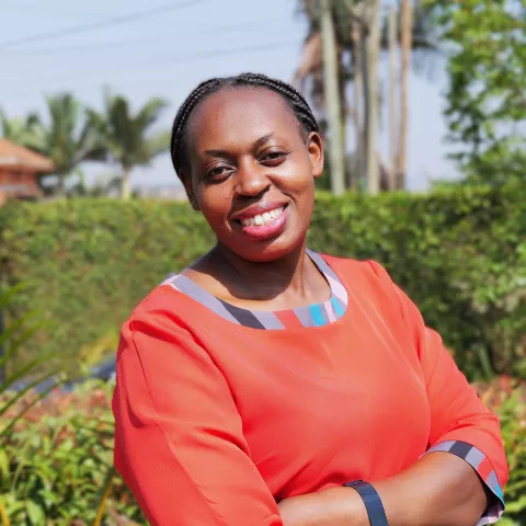 Doreen smiles at the camera. She wears an orange shirt with colourful squares. She is standing in front of green plants. 