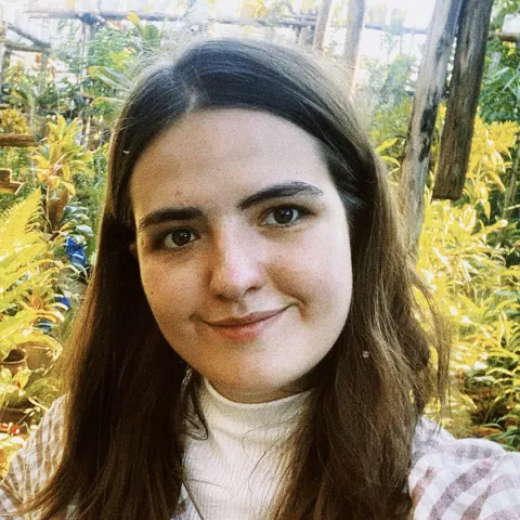 Ellen wears a white shirt and checkered blazer. She is smiling softly. The background has green plants. 