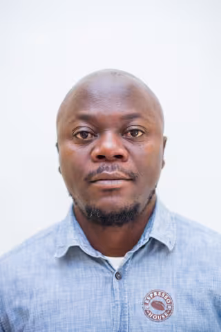 Gondwe is wearing a blue shirt with a coffee bean on it. The background is plain white.