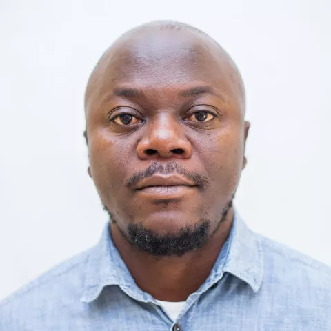 Gondwe is wearing a blue shirt with a coffee bean on it. The background is plain white.