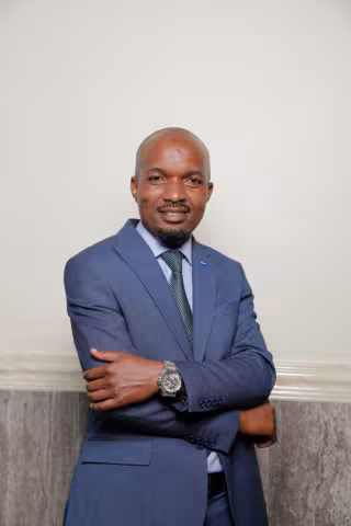 Francis smiles softly with his arms crossed. He wears a blue suit and dark green tie. He is standing in front of a white and grey wall.