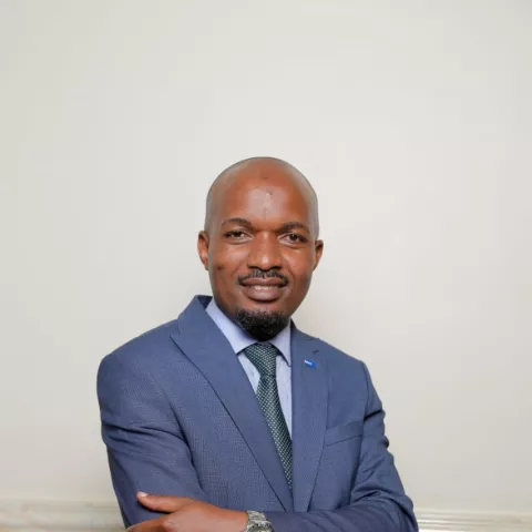 Francis smiles softly with his arms crossed. He wears a blue suit and dark green tie. He is standing in front of a white and grey wall.