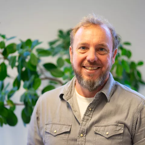 Hans is smiling the camera, wearing a light-brown shirt.