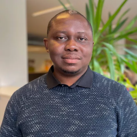 Oyindamola is wearing a dark blue knitted sweater and smiles at the camera. The background is blurred plants.