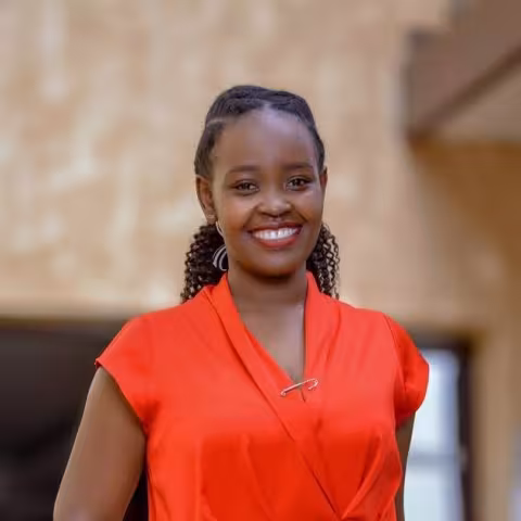 Joanna wears a bright orange shirt. She is smiling softly at the camera. The background is a brown building.