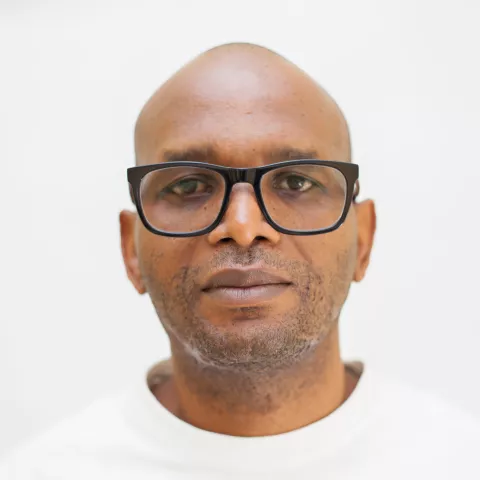 Joseph is wearing glasses and a white top. The background is plain white.