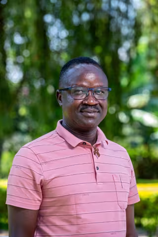 Joseph is wearing a polo shirt and stands outdoors, smiling softly. He's wearing glasses, with a blurred green, leafy background 