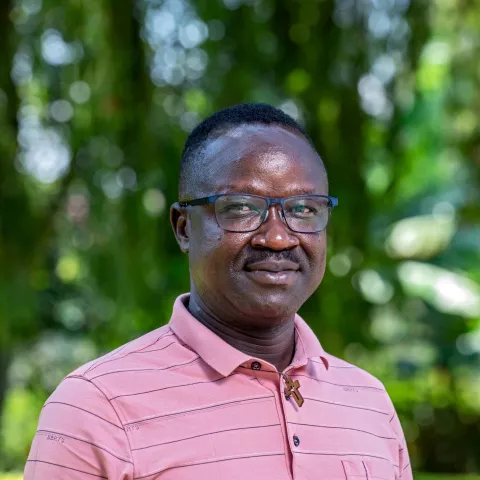 Joseph is wearing a polo shirt and stands outdoors, smiling softly. He's wearing glasses, with a blurred green, leafy background 
