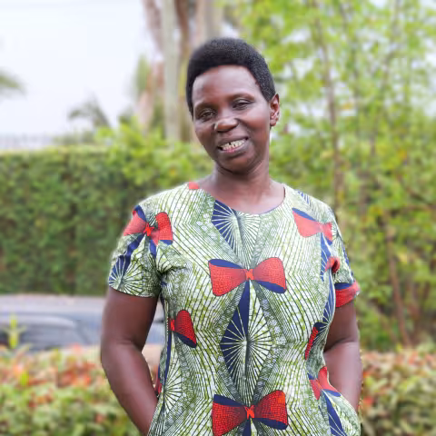 Josephine smiles warmly, wearing a patterned dress with red and blue bows. She stands in front of lush green foliage.