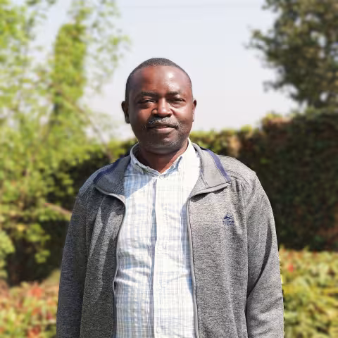 Julius stands outdoors in sunlight, wearing a gray jacket over a checkered shirt. The background is lush greenery.