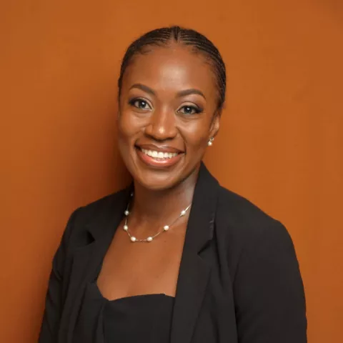 Kakula smiles warmly. She is wearing a black shirt and black blazer and white pearl necklace. She stands in front of a brown backdrop.