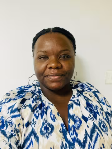 Kalumba is wearing silver earrings and a blue and white patterned top. She is smiling against a white wall. 